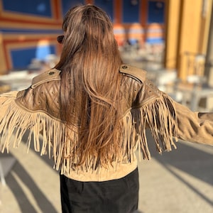 May include: A tan suede jacket with fringe detailing. The jacket has a quilted shoulder design and decorative buttons. The model is wearing a black skirt. The jacket is a stylish piece of outerwear.