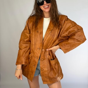 May include: A tan leather blazer with a double-breasted design. The blazer features two front pockets and a classic lapel. The model is wearing sunglasses and denim shorts. The blazer is a medium brown colour.
