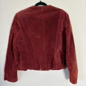 May include: A burgundy suede jacket hangs on a black hanger. The jacket has a round collar, long sleeves, and a zipper closure. The back of the jacket has vertical seams and a slightly flared hemline. The jacket is made of a soft, textured material.