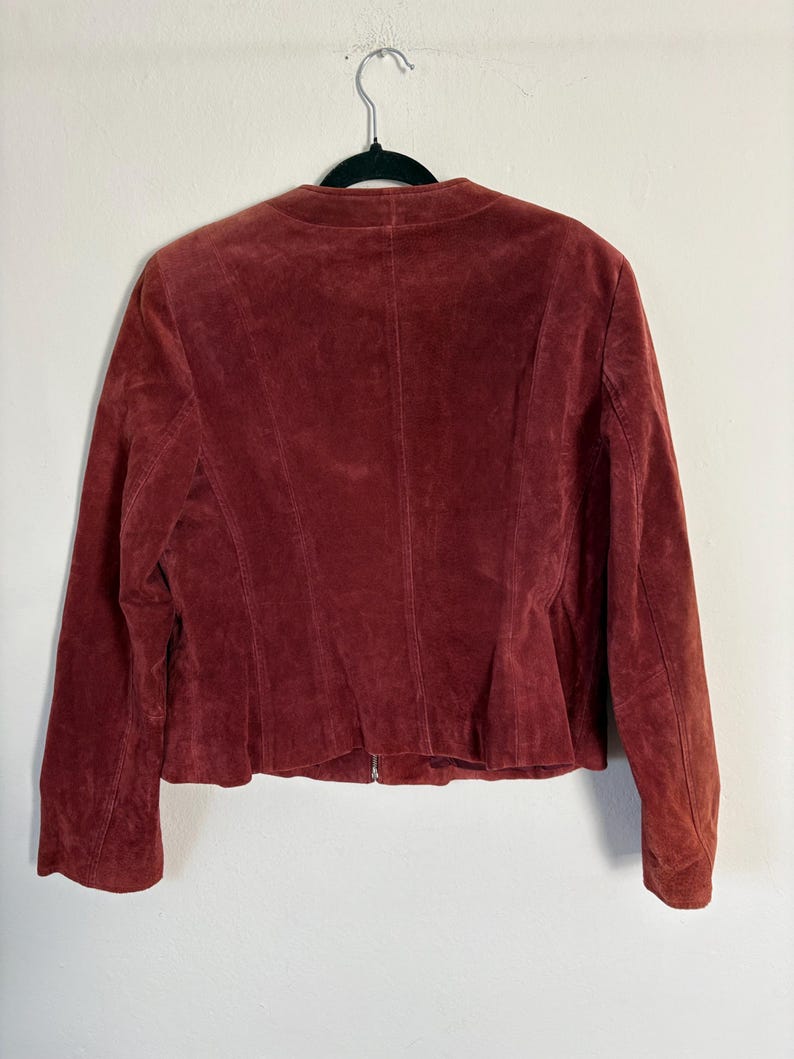 May include: A burgundy suede jacket hangs on a black hanger. The jacket has a round collar, long sleeves, and a zipper closure. The back of the jacket has vertical seams and a slightly flared hemline. The jacket is made of a soft, textured material.