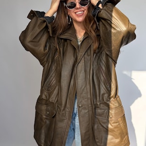 K&ouml;nnte beinhalten: Braune Lederjacke mit mehreren Taschen und Knopfleiste. Die Jacke hat ein klassisches Design mit Kragen und lockerer Passform. Die Jacke wird &uuml;ber einem schwarzen Hemd und einer blauen Jeans getragen. Vintage-Stil.