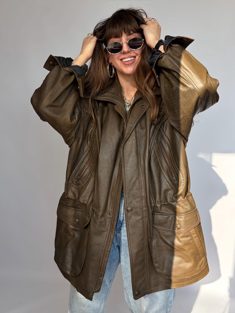 K&ouml;nnte beinhalten: Braune Lederjacke mit mehreren Taschen und Knopfleiste. Die Jacke hat ein klassisches Design mit Kragen und lockerer Passform. Die Jacke wird &uuml;ber einem schwarzen Hemd und einer blauen Jeans getragen. Vintage-Stil.
