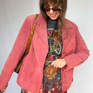 May include: A pink suede blazer with a notched lapel, long sleeves, and two front buttons. It is worn over a black graphic t-shirt with red and yellow text and a dark red skirt featuring a large-scale floral print in orange and green tones.