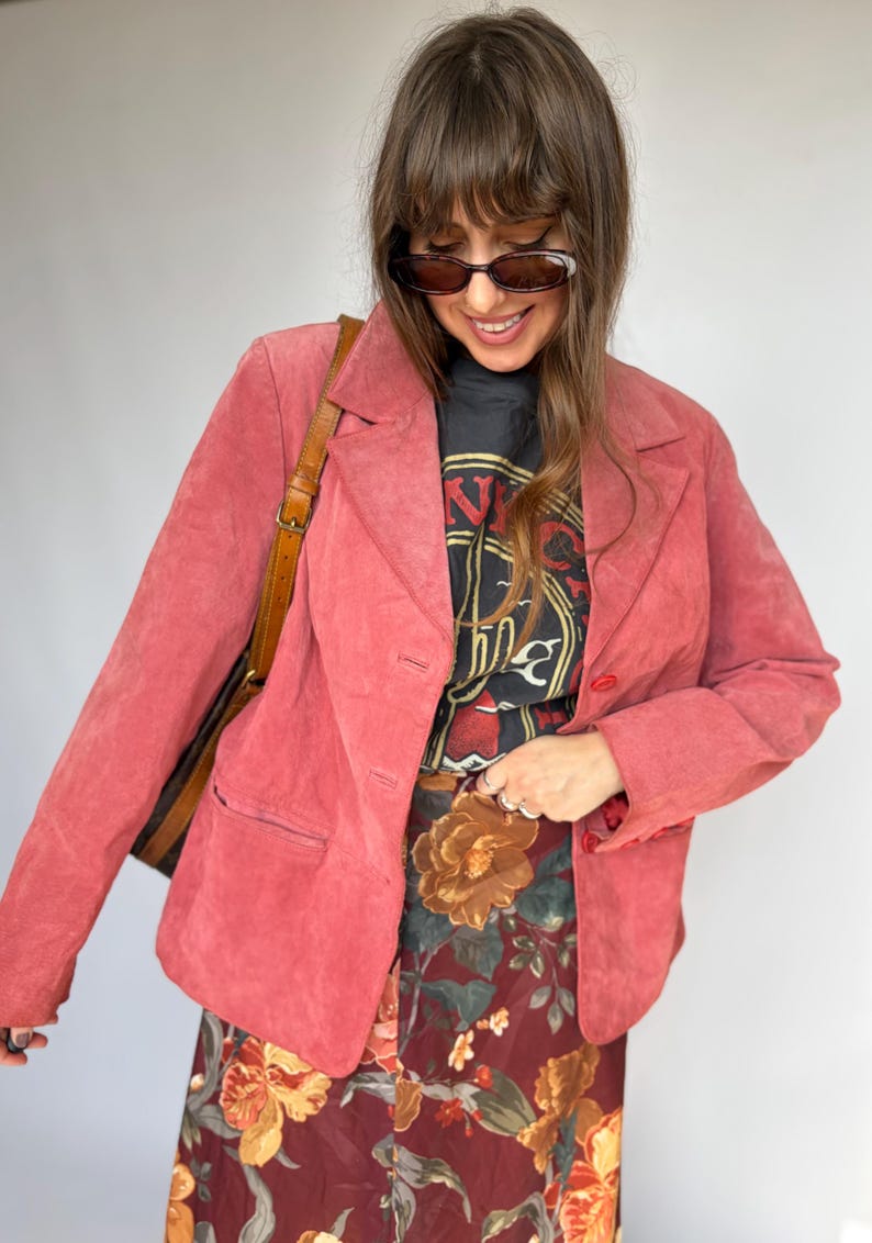 May include: A pink suede blazer with a notched lapel, long sleeves, and two front buttons. It is worn over a black graphic t-shirt with red and yellow text and a dark red skirt featuring a large-scale floral print in orange and green tones.
