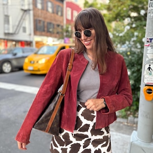 May include: A burgundy suede jacket with a round collar and long sleeves. The person is wearing a gray top and a brown and white cow print skirt. A brown shoulder bag is also visible.