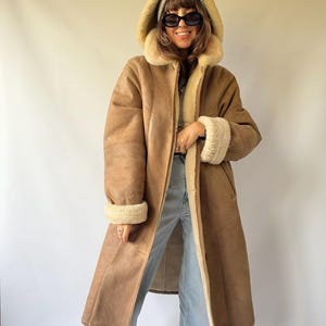 Può includere: Un cappotto lungo color marrone chiaro con fodera in montone color crema, un ampio cappuccio e polsini risvoltati. Il cappotto presenta una texture esterna liscia e una chiusura con bottoni. È indossato sopra jeans azzurri e un berretto in maglia.
