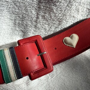 May include: A wide red belt with a large square buckle and a silver-tone metal clasp. The belt features a cream-colored heart applique and striped ribbon detailing in green, white, and navy blue.