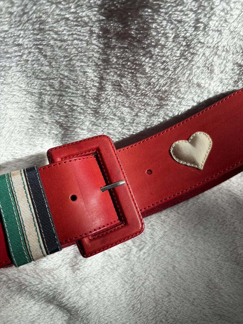 May include: A wide red belt with a large square buckle and a silver-tone metal clasp. The belt features a cream-colored heart applique and striped ribbon detailing in green, white, and navy blue.
