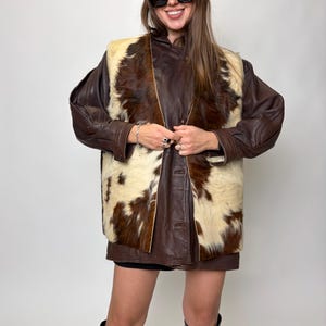 May include: A brown leather jacket with a cowhide vest. The vest is cream and brown. The person is wearing a brown beret and black sunglasses. The person is also wearing black boots.
