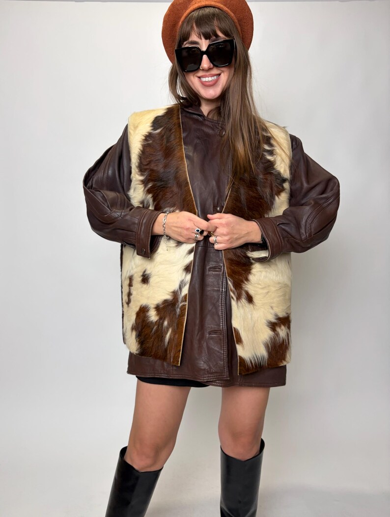 May include: A brown leather jacket with a cowhide vest. The vest is cream and brown. The person is wearing a brown beret and black sunglasses. The person is also wearing black boots.