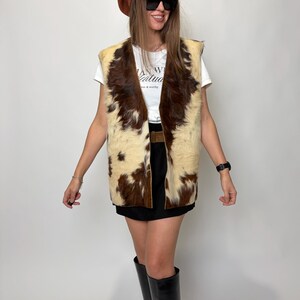 May include: A brown and cream-colored cowhide vest with a V-neck design. The vest is worn over a white t-shirt and black skirt. The person is wearing a brown beret, black sunglasses, and black knee-high boots.