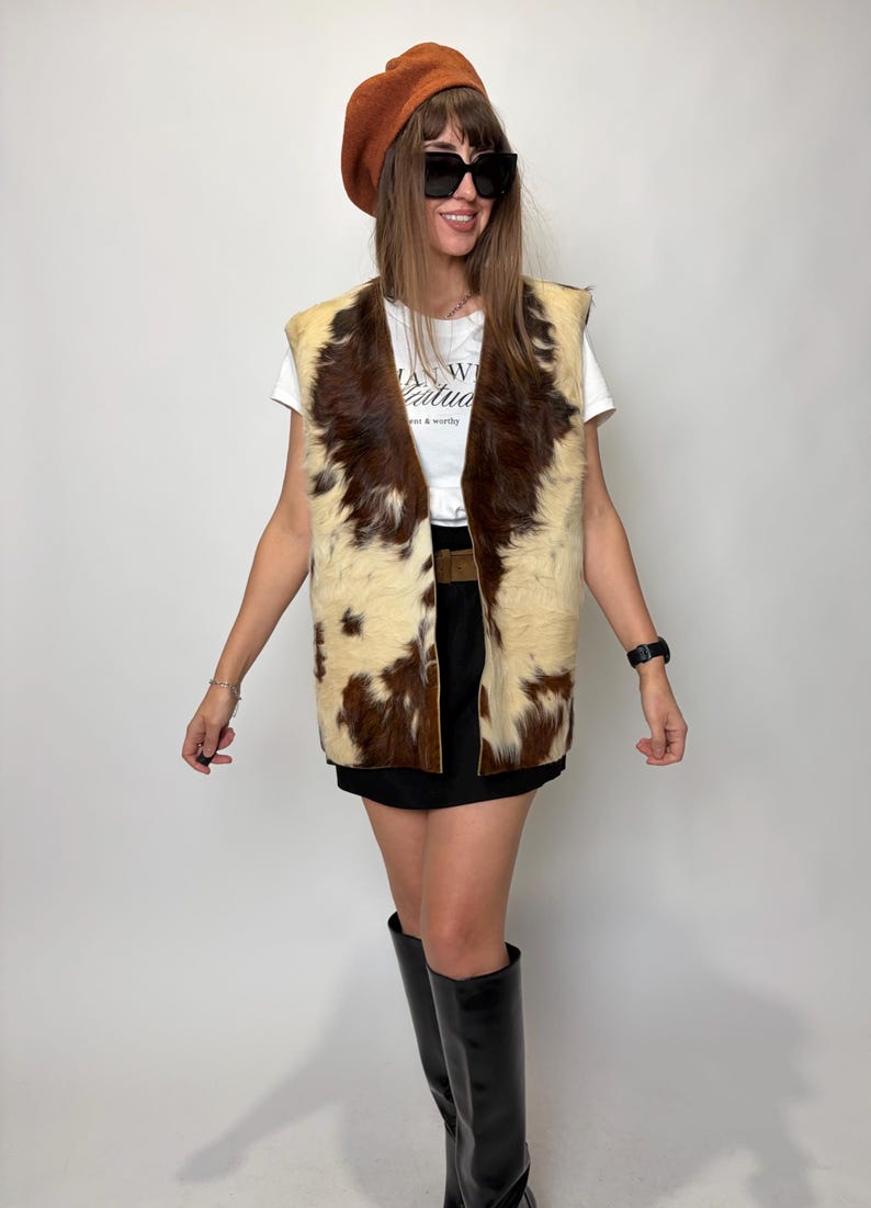 May include: A brown and cream-colored cowhide vest with a V-neck design. The vest is worn over a white t-shirt and black skirt. The person is wearing a brown beret, black sunglasses, and black knee-high boots.