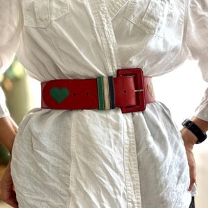 May include: A red leather belt with a rectangular buckle. The belt features a green heart and a gold heart design. A section of the belt has green, navy, and white stripes. The belt is worn over a white shirt.