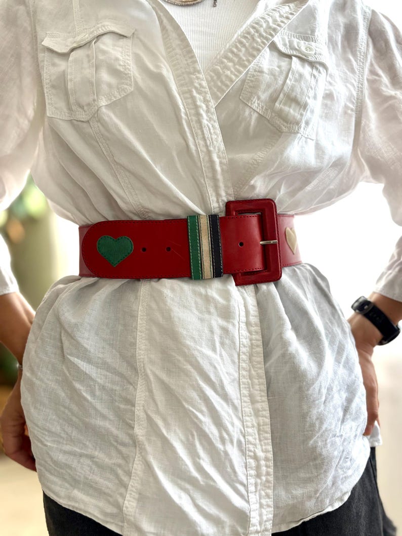 May include: A red leather belt with a rectangular buckle. The belt features a green heart and a gold heart design. A section of the belt has green, navy, and white stripes. The belt is worn over a white shirt.