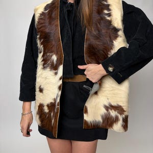 May include: A cowhide vest with a brown and cream pattern. The vest is sleeveless and open in the front. The model is wearing a black long-sleeve shirt and a black skirt.
