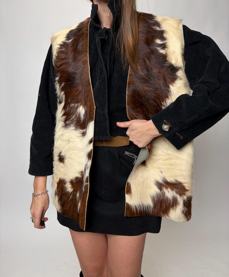 May include: A cowhide vest with a brown and cream pattern. The vest is sleeveless and open in the front. The model is wearing a black long-sleeve shirt and a black skirt.
