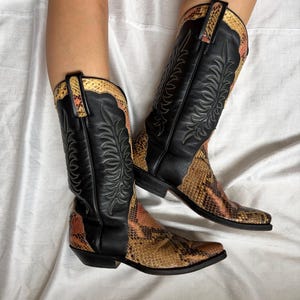 Puede incluir: Botas de vaquero de cuero negro con detalles de piel de serpiente. Las botas tienen punta afilada y costuras decorativas. El estampado de piel de serpiente presenta tonos marrones, naranjas y beige.