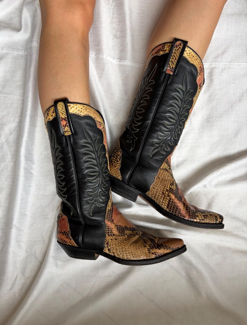 Puede incluir: Botas de vaquero de cuero negro con detalles de piel de serpiente. Las botas tienen punta afilada y costuras decorativas. El estampado de piel de serpiente presenta tonos marrones, naranjas y beige.