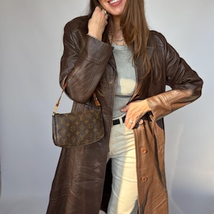 May include: A brown leather trench coat with button closures, paired with a small brown handbag. The outfit includes a gray top, cream-colored pants, and black sunglasses. The handbag has a brown monogram pattern.