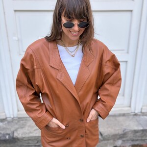 May include: A tan leather blazer featuring a double-breasted front with two visible buttons, wide lapels, and long sleeves. The jacket is worn over a plain white crew-neck t-shirt, paired with light blue denim jeans and oval-shaped sunglasses.