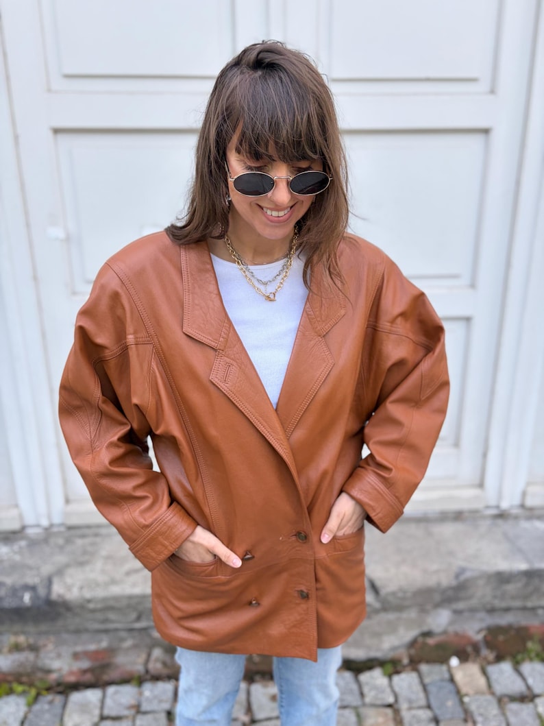 May include: A tan leather blazer featuring a double-breasted front with two visible buttons, wide lapels, and long sleeves. The jacket is worn over a plain white crew-neck t-shirt, paired with light blue denim jeans and oval-shaped sunglasses.