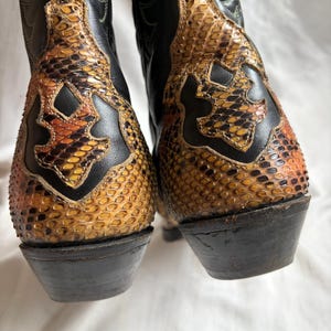 Puede incluir: Un par de botas de vaquero de cuero negro con detalles de piel de serpiente en tonos marr&oacute;n, naranja y amarillo. Las botas tienen puntas afiladas y tacones apilados, un estilo Western cl&aacute;sico.