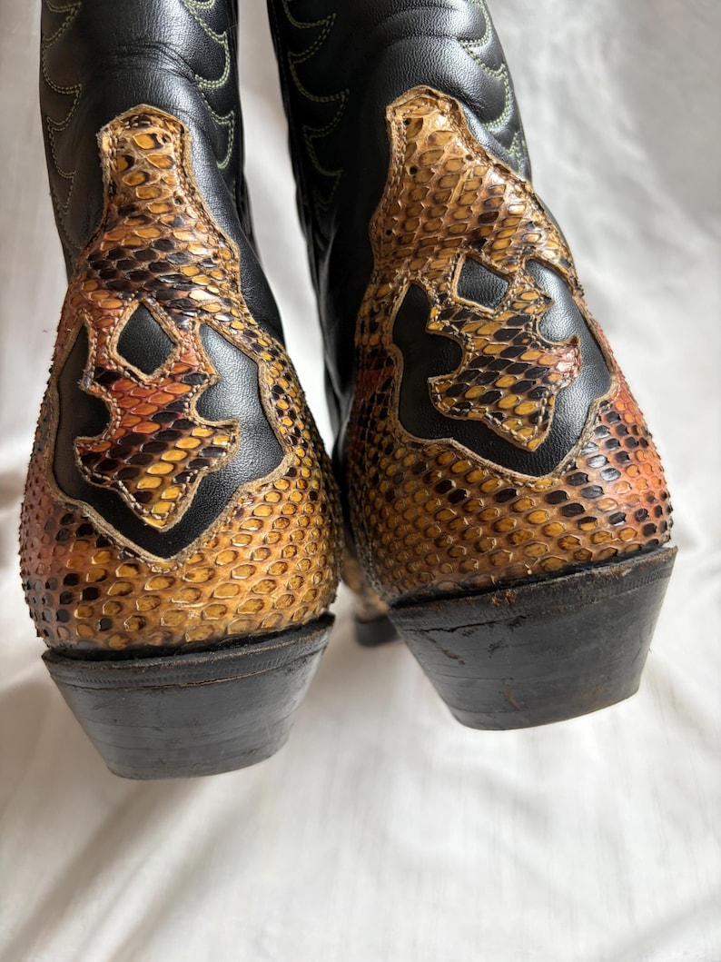 Puede incluir: Un par de botas de vaquero de cuero negro con detalles de piel de serpiente en tonos marr&oacute;n, naranja y amarillo. Las botas tienen puntas afiladas y tacones apilados, un estilo Western cl&aacute;sico.