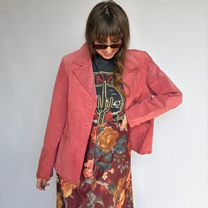 May include: A dusty rose blazer with a three-button closure, paired with a black graphic tee and a floral print skirt. The skirt features a burgundy background with large orange and yellow flowers. The tee has a desert cactus graphic.