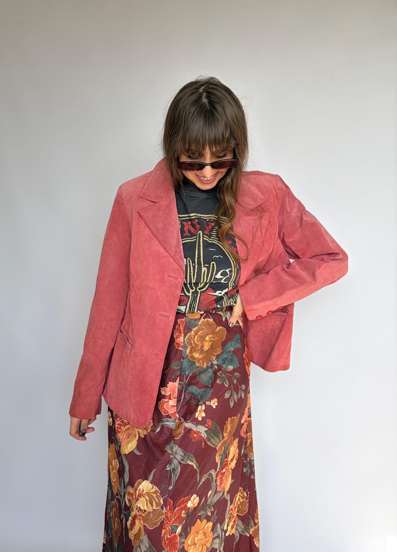 May include: A dusty rose blazer with a three-button closure, paired with a black graphic tee and a floral print skirt. The skirt features a burgundy background with large orange and yellow flowers. The tee has a desert cactus graphic.