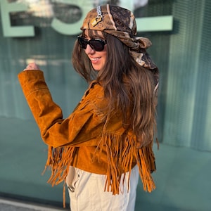May include: A tobacco-colored suede jacket featuring long fringe detailing along the back and sleeves, paired with a brown patterned headscarf adorned with a small gold-toned padlock charm and black oversized sunglasses.