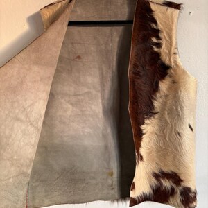May include: A sleeveless vest made of cowhide, featuring a mix of brown and cream-colored fur on the exterior. The interior is a light brown suede-like material. The vest hangs on a black hanger against a white wall.