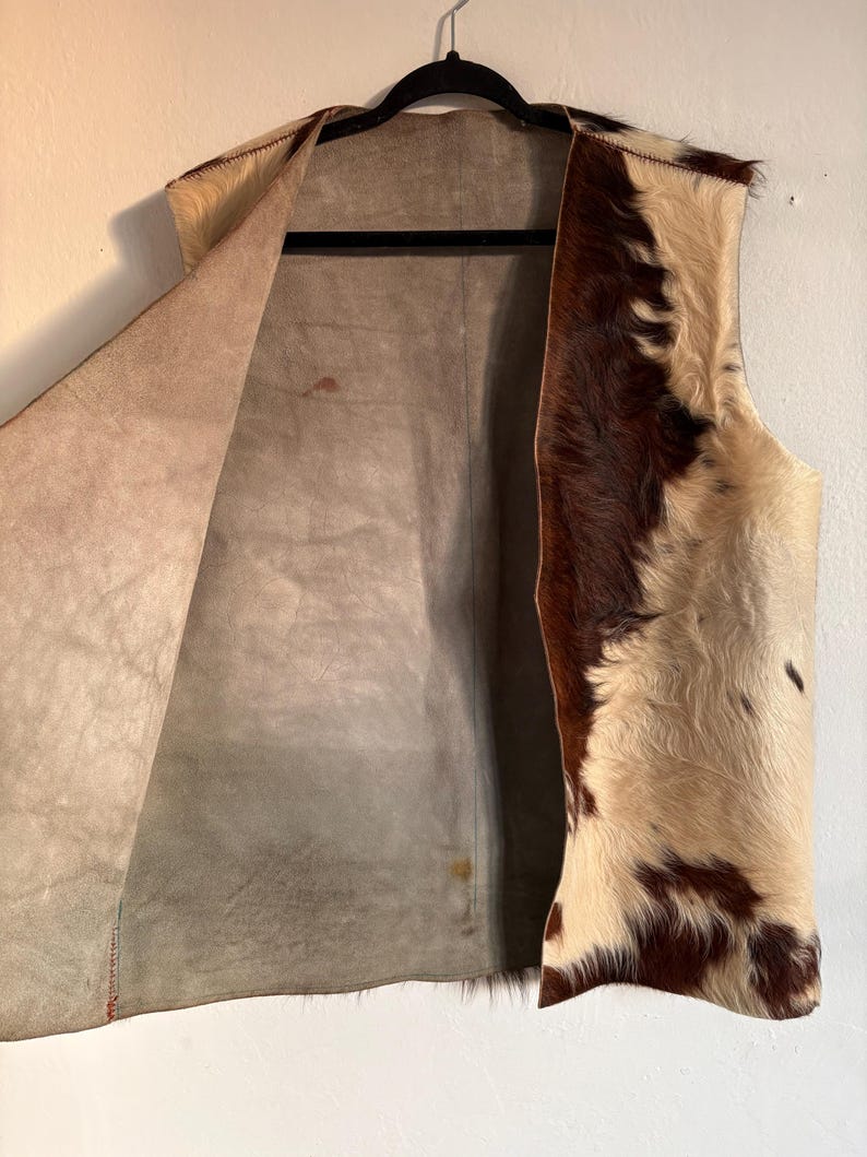 May include: A sleeveless vest made of cowhide, featuring a mix of brown and cream-colored fur on the exterior. The interior is a light brown suede-like material. The vest hangs on a black hanger against a white wall.