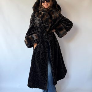 May include: A long, black, textured faux fur coat featuring a large hood with brown faux fur trim, wide sleeves with matching brown faux fur cuffs, and a deep, patterned pile throughout the body. The coat is worn over blue denim jeans.