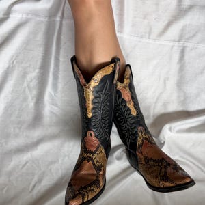 Puede incluir: Botas de vaquero negras con empeines y detalles con estampado de piel de serpiente. Las botas tienen una puntera puntiaguda y costuras decorativas en la ca&ntilde;a. El estampado de piel de serpiente presenta tonos marrones, beiges y naranjas.
