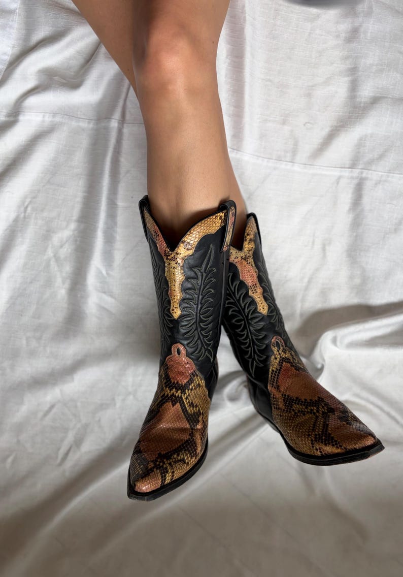 Puede incluir: Botas de vaquero negras con empeines y detalles con estampado de piel de serpiente. Las botas tienen una puntera puntiaguda y costuras decorativas en la ca&ntilde;a. El estampado de piel de serpiente presenta tonos marrones, beiges y naranjas.