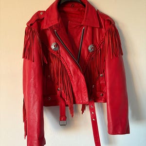 Op de afbeelding: Een felrode leren jas met franjes op de mouwen en borst. De jas heeft een klassieke bikerstijl met een reverskraag, een diagonale rits en zilverkleurige metalen knopen en accenten. Een riem met gesp is inbegrepen.