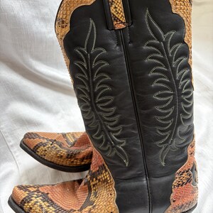 Puede incluir: Botas de vaquero con ca&ntilde;a de cuero negro y estampado de piel de serpiente en el pie y el borde superior. Las botas tienen costuras decorativas y una puntera puntiaguda. El estampado de piel de serpiente presenta tonos naranja, marr&oacute;n y amarillo.