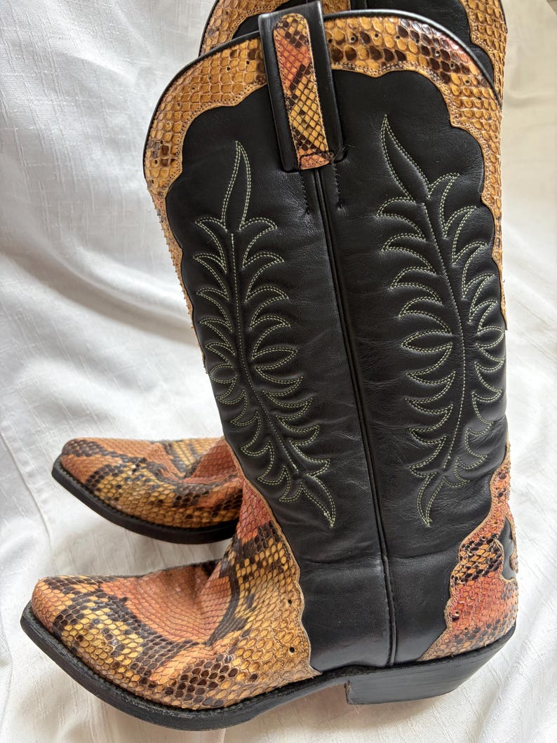 Puede incluir: Botas de vaquero con ca&ntilde;a de cuero negro y estampado de piel de serpiente en el pie y el borde superior. Las botas tienen costuras decorativas y una puntera puntiaguda. El estampado de piel de serpiente presenta tonos naranja, marr&oacute;n y amarillo.