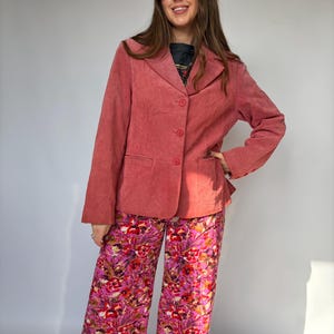 May include: A dusty rose blazer with three buttons, paired with wide-leg pants featuring a vibrant floral pattern in shades of pink, red, and white. The outfit is a stylish ensemble.