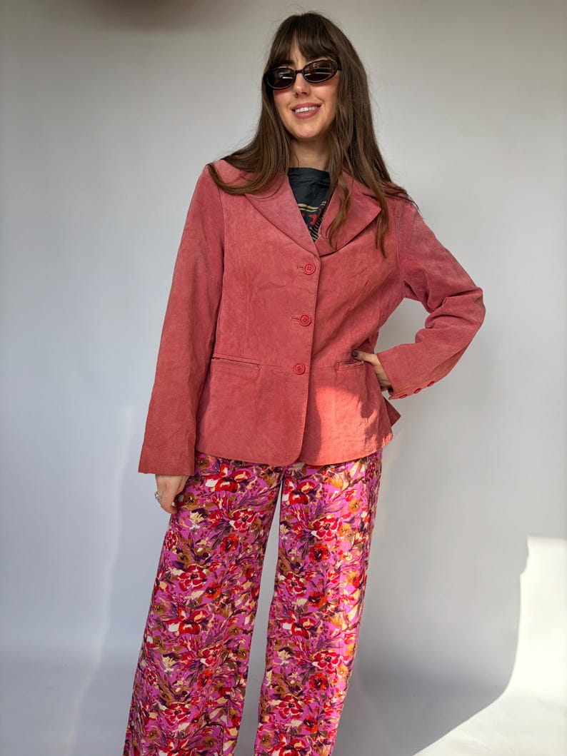 May include: A dusty rose blazer with three buttons, paired with wide-leg pants featuring a vibrant floral pattern in shades of pink, red, and white. The outfit is a stylish ensemble.