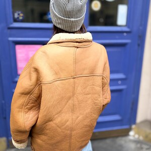 May include: A tan shearling jacket with a cream-colored shearling lining and a gray ribbed knit beanie. The jacket has a classic design with visible stitching and a wide collar. The person is wearing light blue jeans.