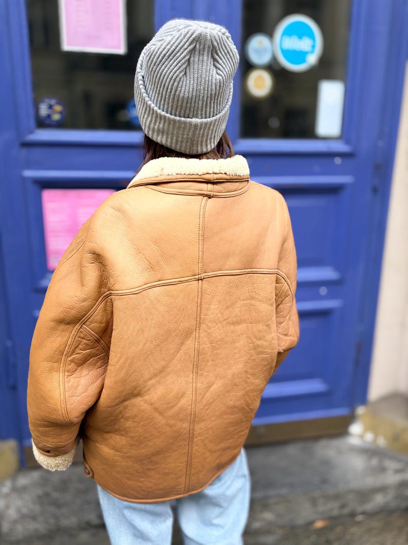 May include: A tan shearling jacket with a cream-colored shearling lining and a gray ribbed knit beanie. The jacket has a classic design with visible stitching and a wide collar. The person is wearing light blue jeans.