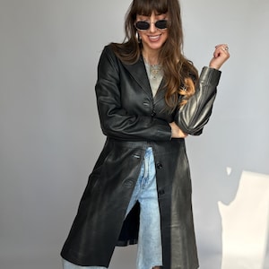 May include: A black leather coat with a button-up front and a long, flowing design. The coat is knee-length and has a classic, tailored look. The coat is worn over light-wash denim jeans.