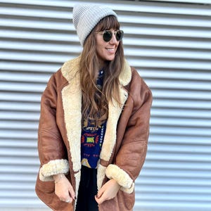 Puede incluir: Un gorro de punto acanalado gris claro, gafas de sol redondas oscuras y una chaqueta de cuero marrón con un grueso forro de piel de oveja color crema. Debajo, un suéter azul oscuro con un patrón geométrico colorido y pantalones negros.