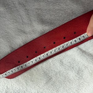 May include: A red leather belt with a series of evenly spaced holes. A measuring tape is placed along the belt, showing measurements from 67 to 89 inches. The belt is on a white, textured surface.