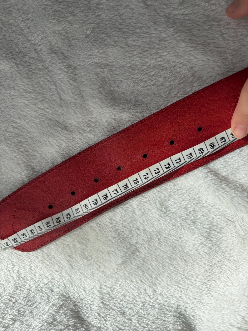 May include: A red leather belt with a series of evenly spaced holes. A measuring tape is placed along the belt, showing measurements from 67 to 89 inches. The belt is on a white, textured surface.