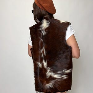 May include: A brown and white fur vest with a unique pattern. The vest is made of animal fur and has a dark brown base color with white markings. The person in the image is wearing a brown beret and a white t-shirt.