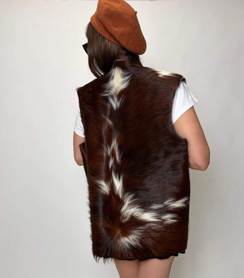 May include: A brown and white fur vest with a unique pattern. The vest is made of animal fur and has a dark brown base color with white markings. The person in the image is wearing a brown beret and a white t-shirt.