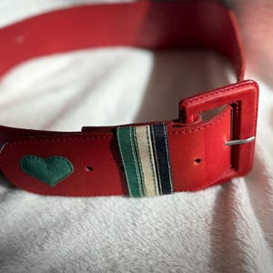 May include: A red belt with a rectangular buckle and a green heart-shaped patch. The belt features a striped accent with green, white, and navy blue stripes. The belt is made of a smooth material.