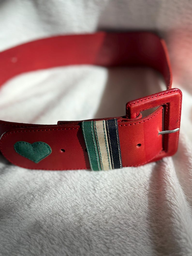 May include: A red belt with a rectangular buckle and a green heart-shaped patch. The belt features a striped accent with green, white, and navy blue stripes. The belt is made of a smooth material.